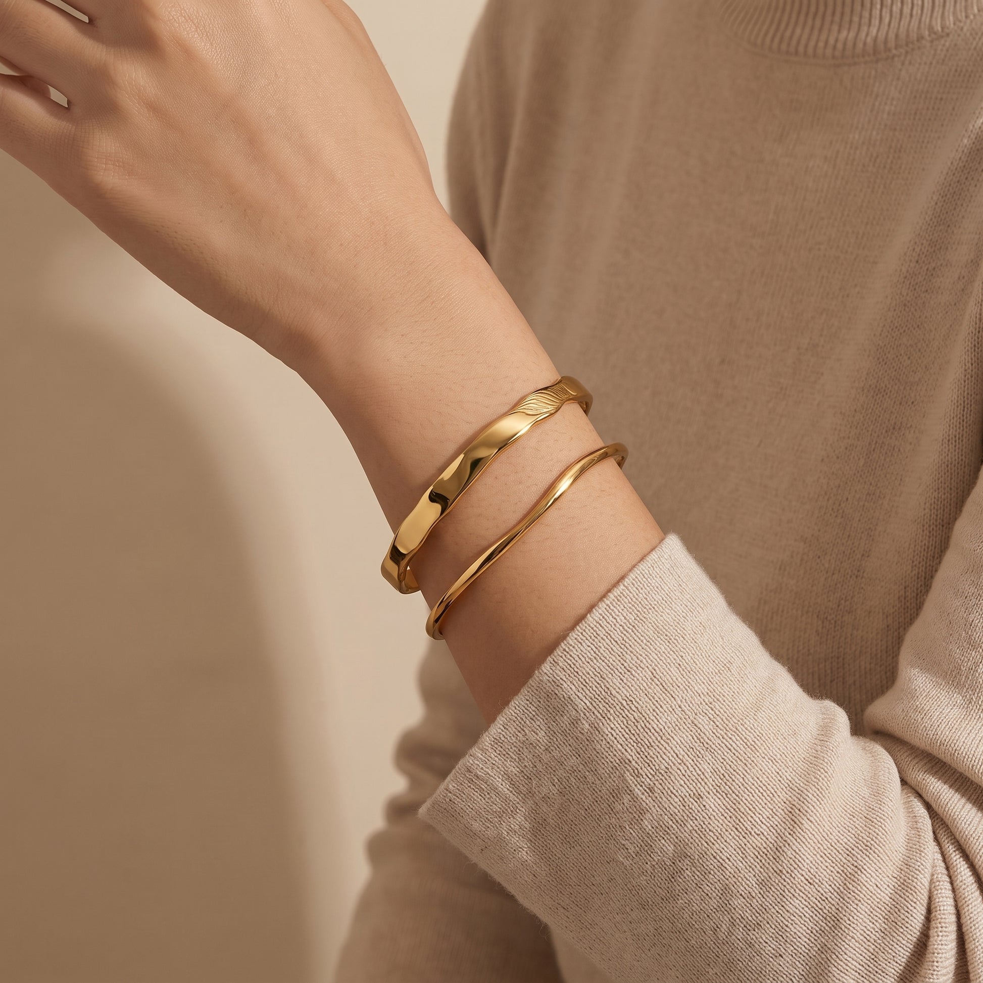 Modern gold wrist stack on a woman featuring the skin friendly Ondine slim wave cuff layered with the statement wave bangle against a beige knit sweater.