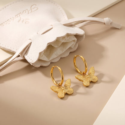 Pair of waterproof Papillon gold butterfly charm hoop earrings displayed next to a cream Floravisha scalloped velvet pouch.