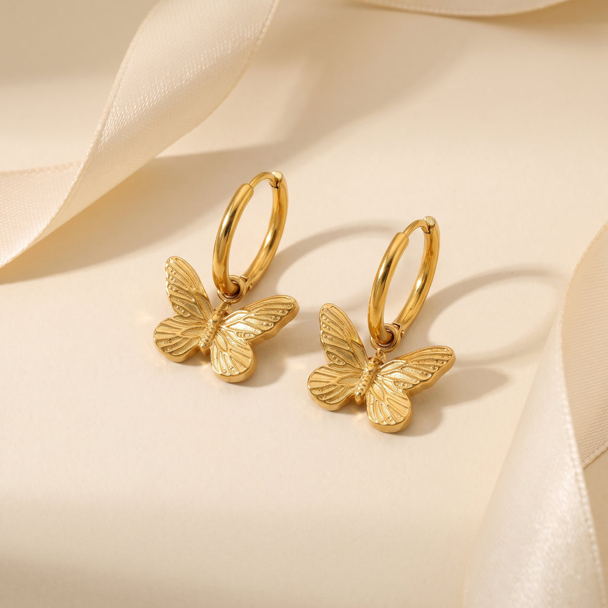 High quality Papillon gold butterfly huggie hoop earrings showcasing etched textured wings on a beige surface with ivory ribbon.