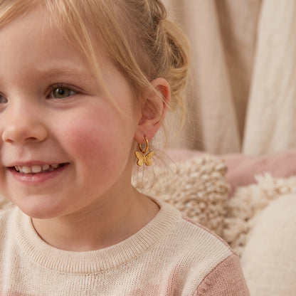 Close-up of a smiling young girl wearing the hypoallergenic Papillon gold butterfly charm huggie earrings with a cream sweater.