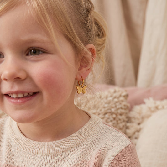 Close-up of a smiling young girl wearing the hypoallergenic Papillon gold butterfly charm huggie earrings with a cream sweater.
