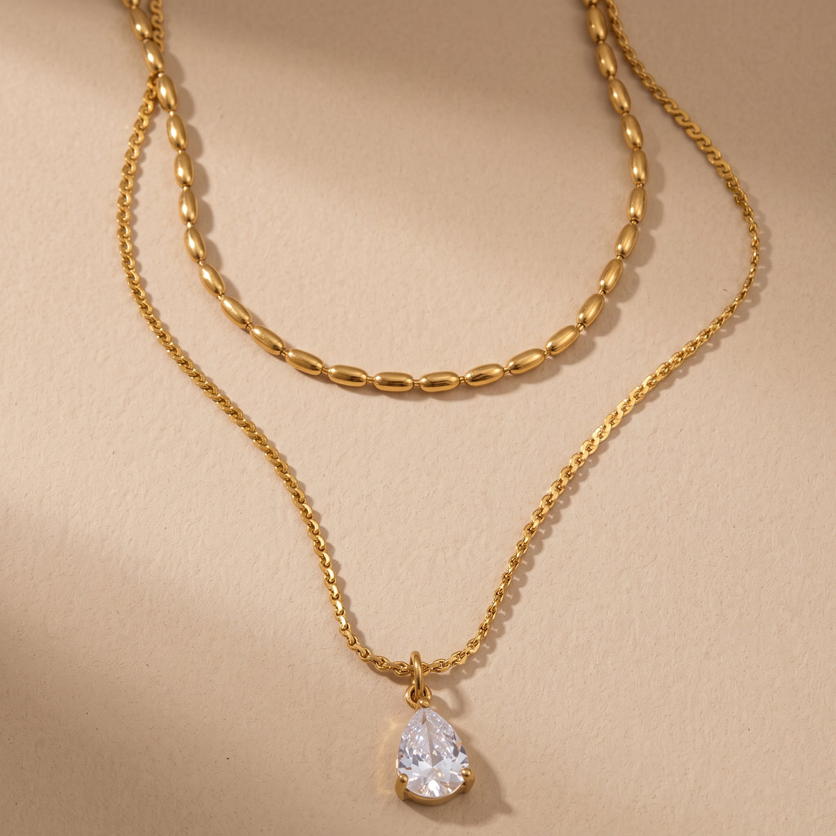 Tarnish-free Seraphine gold layered necklace displaying an upper oval bead chain and lower pear-cut crystal teardrop pendant on a smooth beige background.