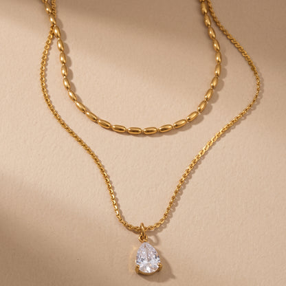 Tarnish-free Seraphine gold layered necklace displaying an upper oval bead chain and lower pear-cut crystal teardrop pendant on a smooth beige background.