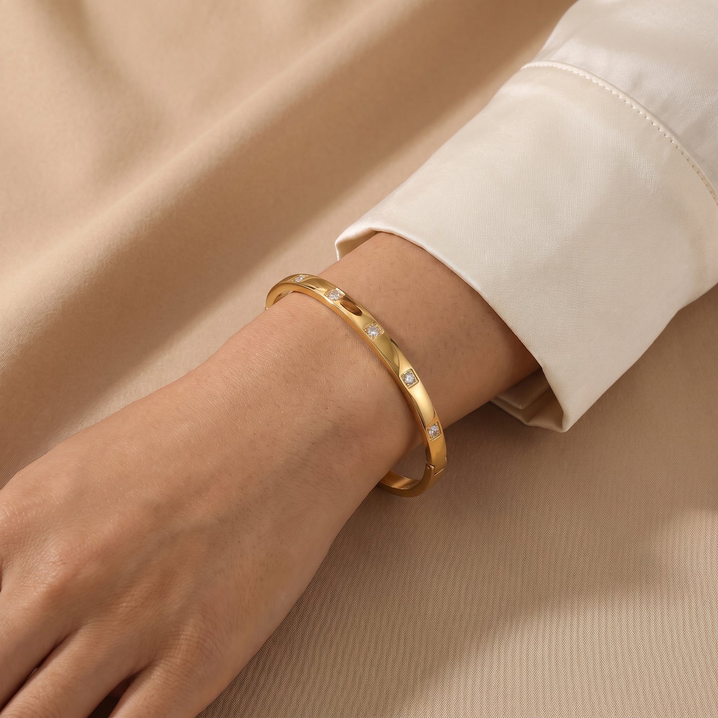 Close-up view of a woman's wrist wearing the skin friendly Solene gold flush-set crystal oval bracelet with a cream silk shirt.