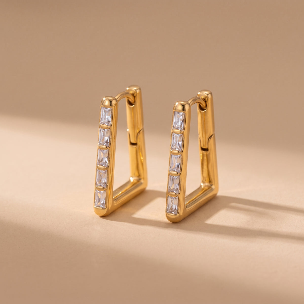 High quality Vendôme gold geometric trapezoid hoop earrings featuring channel-set clear baguette crystals resting on a warm beige studio background.