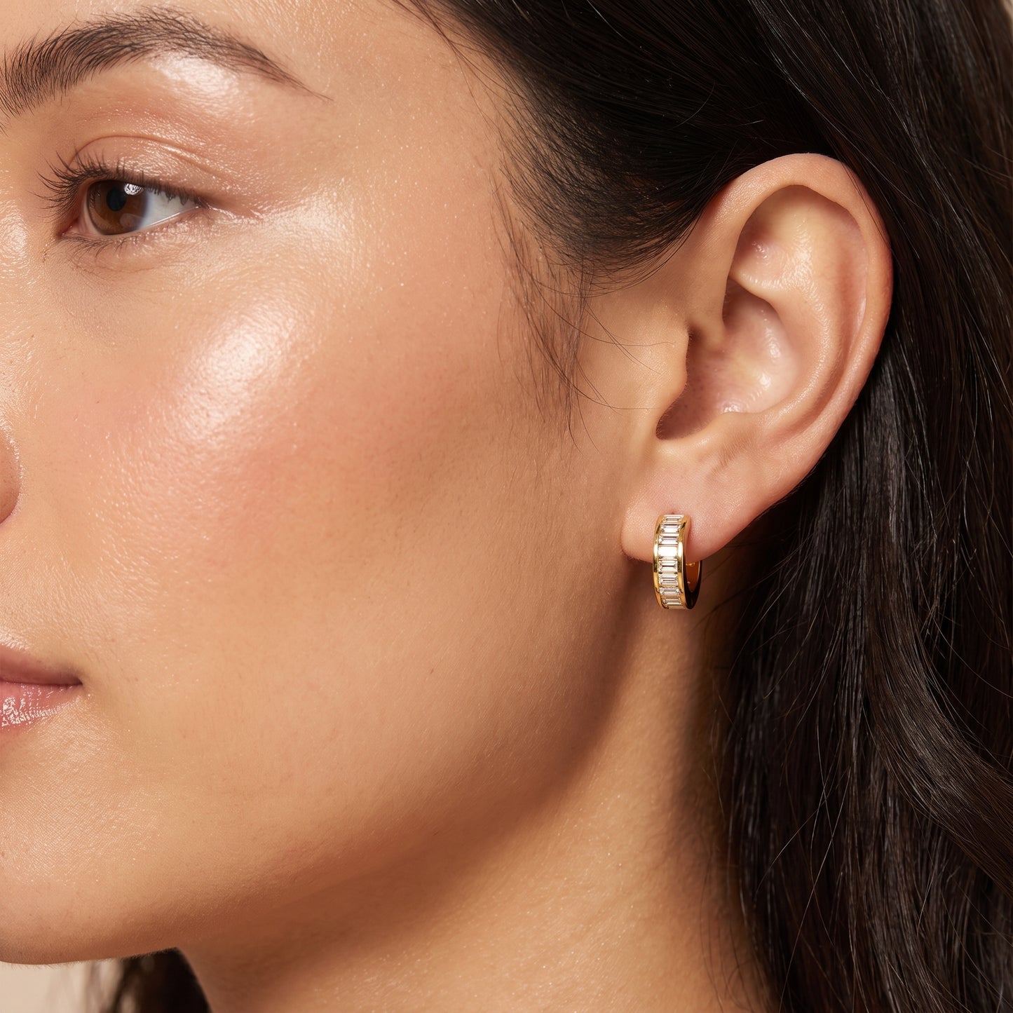 Close-up portrait of a woman wearing the waterproof Verona gold baguette crystal huggie hoop earring on her earlobe.