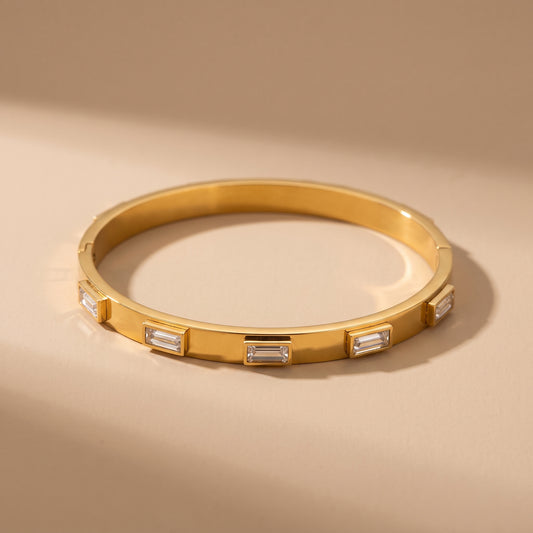High quality Verona Gold Bracelet by Floravisha featuring raised bezel-set clear baguette crystals resting on a warm beige travertine surface under natural sunlight.