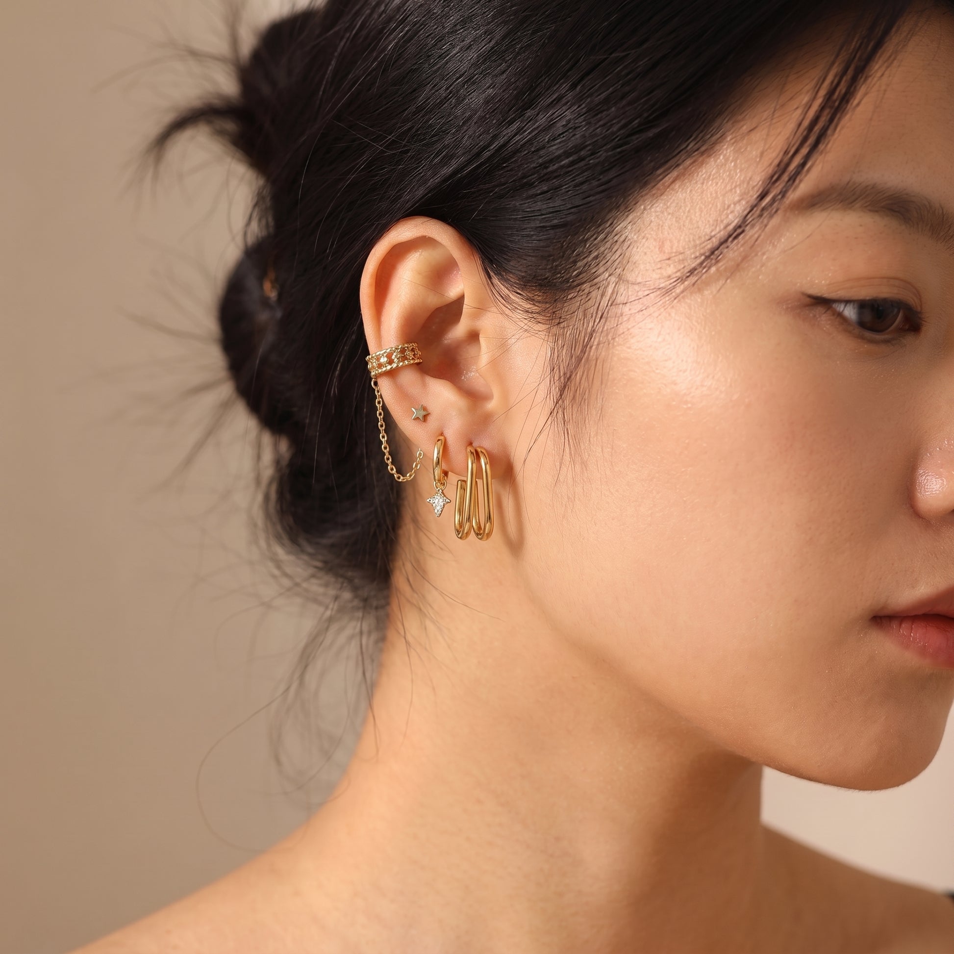 High quality Vienne gold double oval hoop earrings styled in a modern ear stack with a chain ear cuff and star studs on a model's earlobe.
