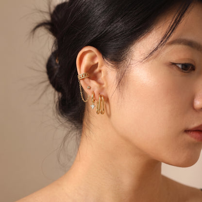 High quality Vienne gold double oval hoop earrings styled in a modern ear stack with a chain ear cuff and star studs on a model's earlobe.