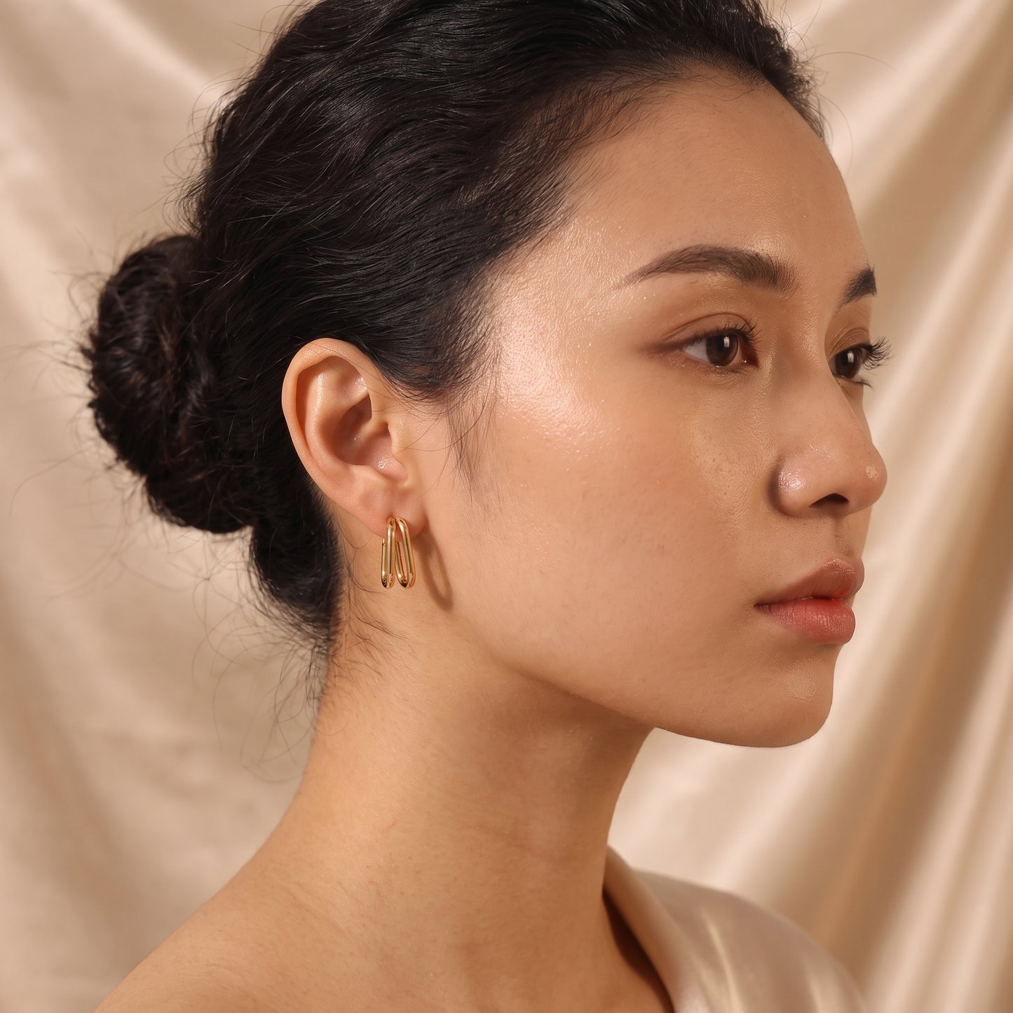 Skin friendly Vienne gold double oval hoop earrings on a model's earlobe set against draped champagne silk in warm natural lighting.